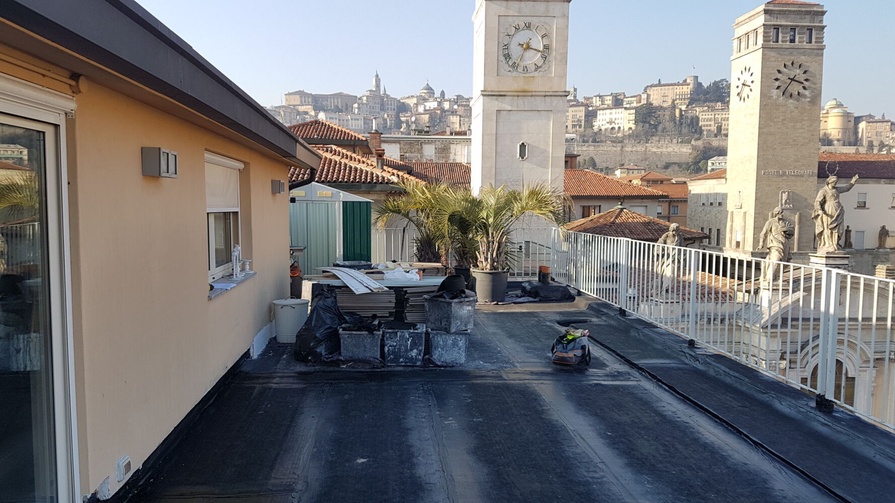Terrazza con guaina bituminosa nera, ringhiera bianca e palme in vaso, torri con orologio e Città Alta di Bergamo